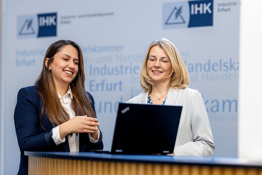 Zwei Frauen im Anzug stehen an einem Tresen und schauen lachend in einen Laptop. Im Hintergrund das IHK Erfurt Logo.
